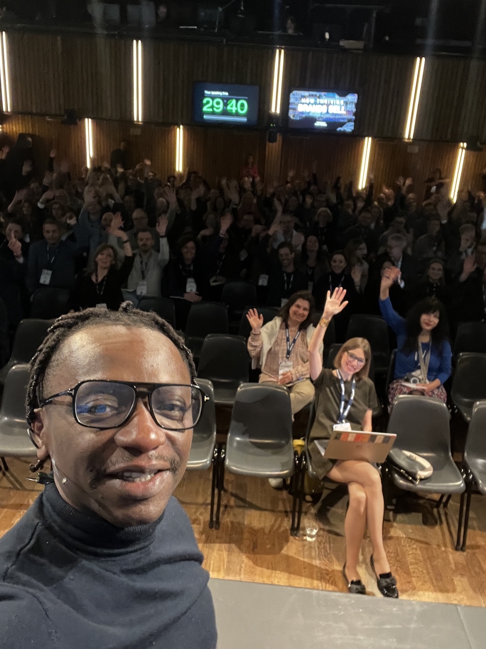 Kunle Campbell taking a photograph with an audience waving behind him in an auditorium.